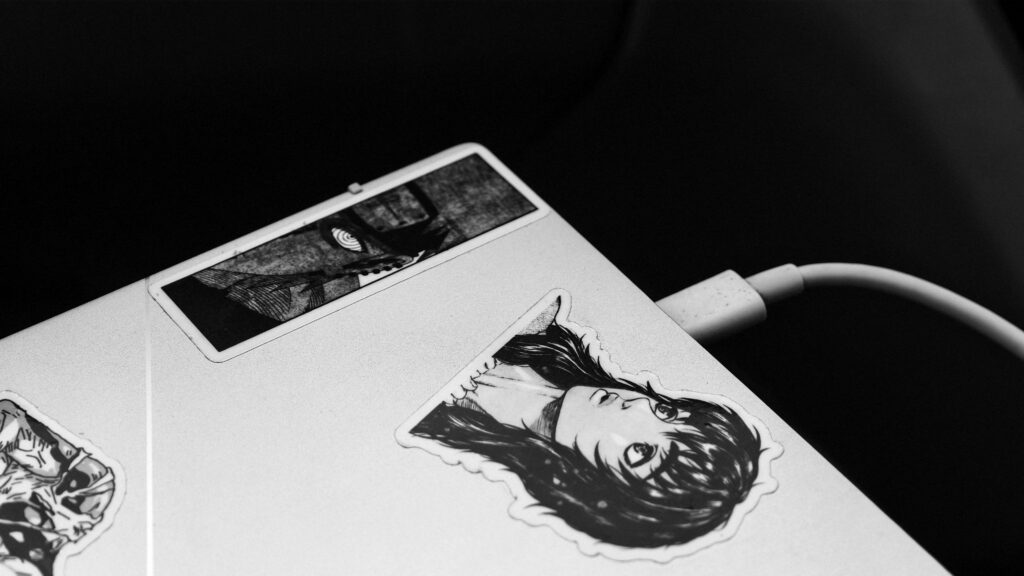 Monochrome image of a laptop adorned with anime-themed stickers and connected to a charger.